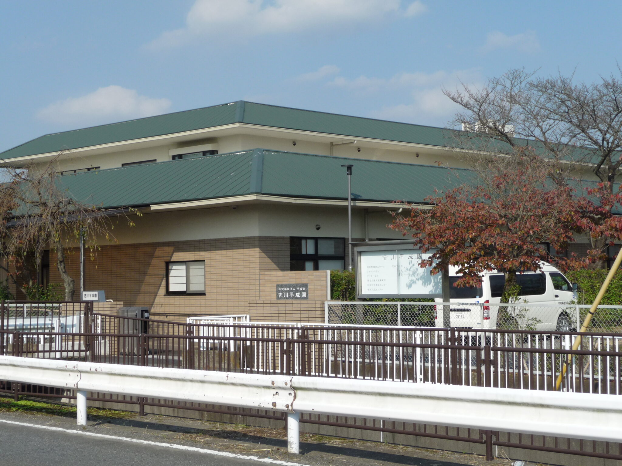 緑色の屋根を持つ平屋造りの介護施設の外観。フェンスと駐車場が見える。